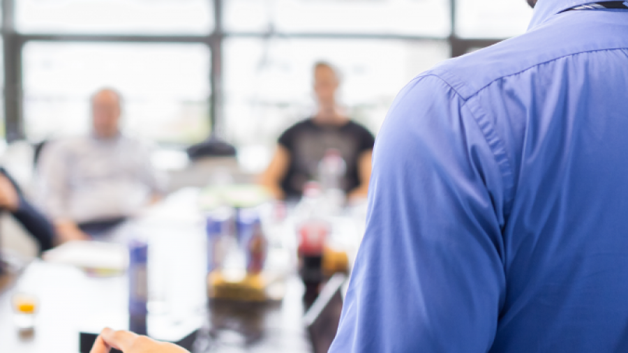 A man in a blue shirt stands and gestures while giving a presentation to a group of people seated around a table in a bright, modern office setting.