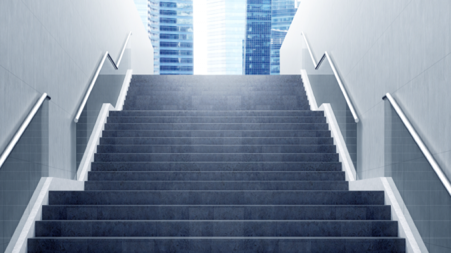 A wide staircase with metal railings on both sides leads upwards towards bright daylight, with modern glass skyscrapers visible in the background.