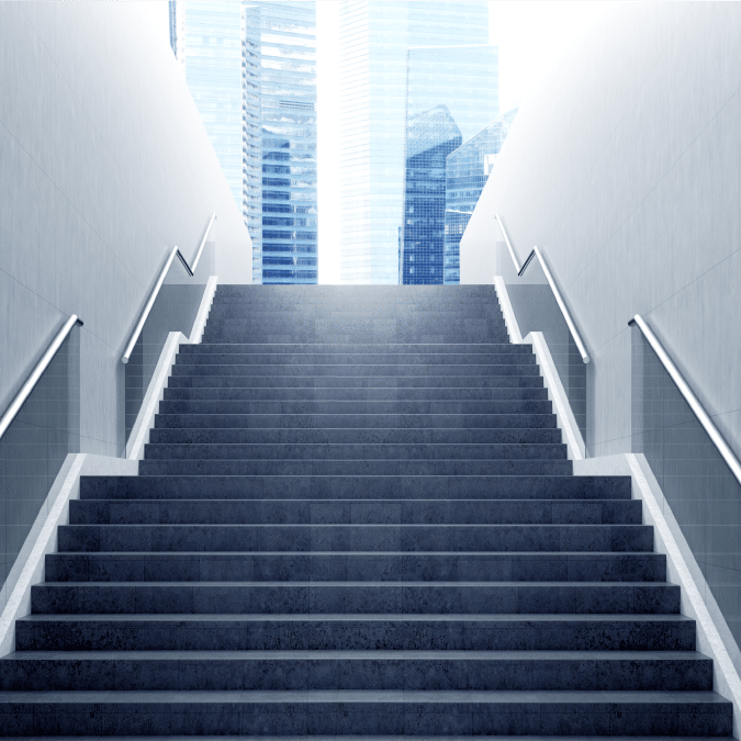 A wide staircase with metal railings on both sides leads upwards towards bright daylight, with modern glass skyscrapers visible in the background.