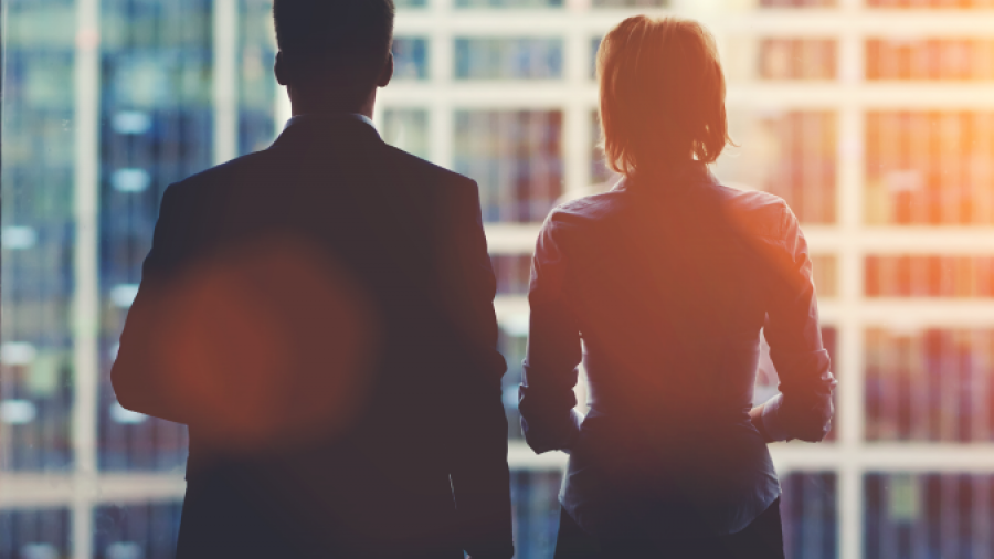 Two business professionals, a man and a woman, are seen from behind standing in front of a large window overlooking tall office buildings in a city, with sunlight streaming in.