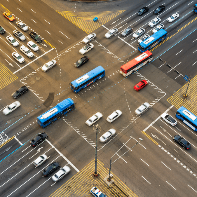 Aerial view of a busy city intersection with multiple lanes of cars, blue buses, and a red car moving in different directions, surrounded by crosswalks and yellow traffic markings.