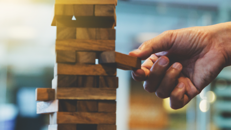 A hand carefully pulls a wooden block from a stacked Jenga tower on a table, with blurred indoor background and warm lighting.