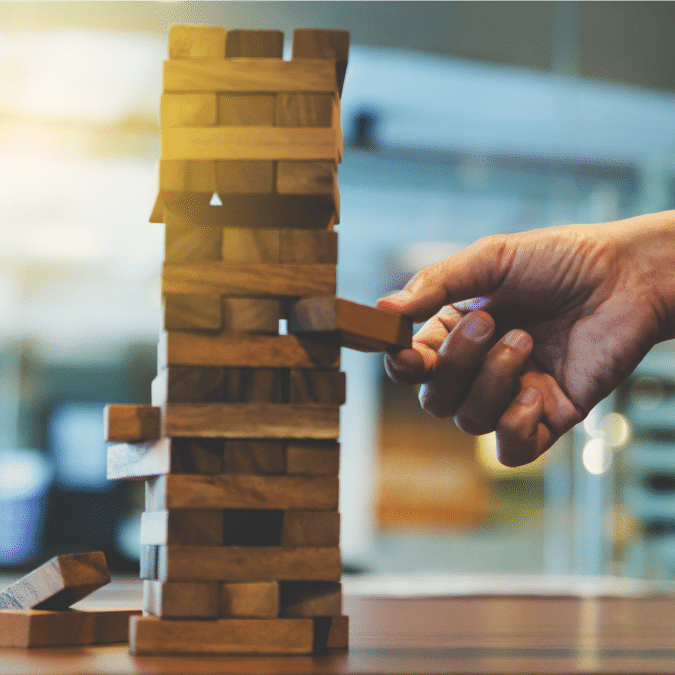 A hand carefully pulls a wooden block from a stacked Jenga tower on a table, with blurred indoor background and warm lighting.