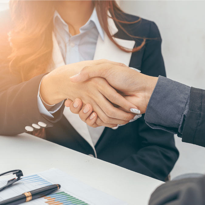 Two people in business attire shake hands over a desk with charts, a pen, and eyeglasses, suggesting a business agreement or successful meeting.