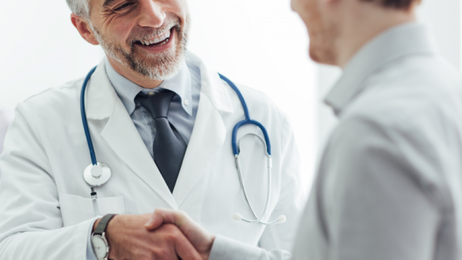 A smiling doctor in a white coat and stethoscope shakes hands with a patient in an office setting, creating a friendly and welcoming atmosphere.