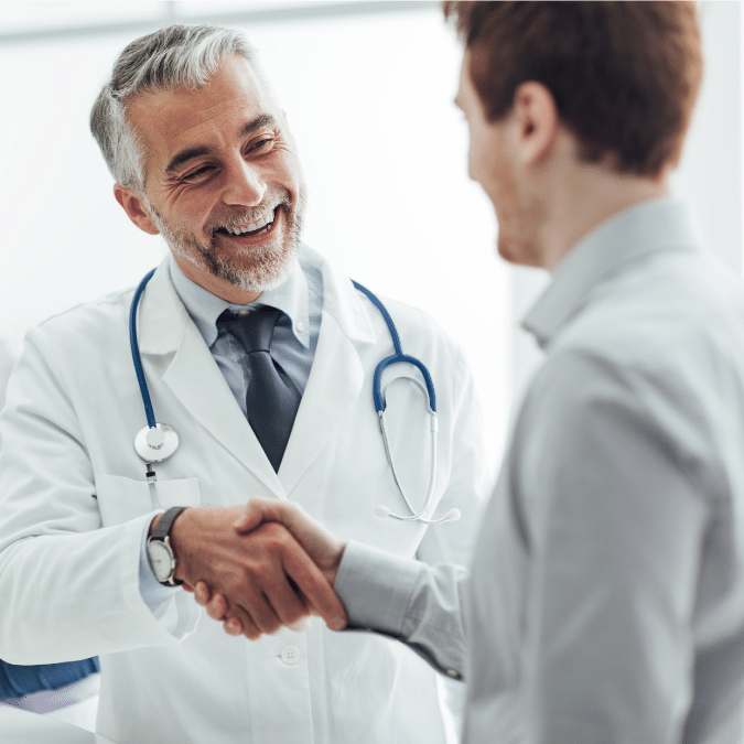 A smiling doctor in a white coat and stethoscope shakes hands with a patient in an office setting, creating a friendly and welcoming atmosphere.