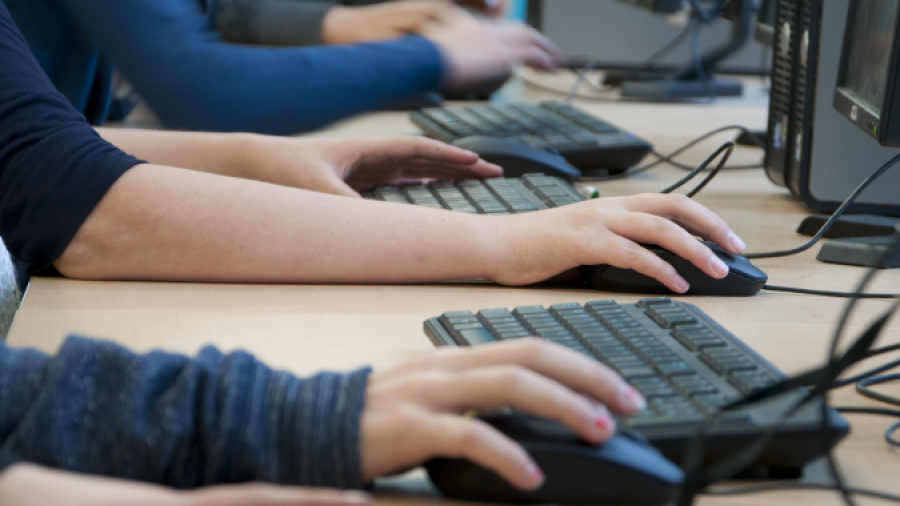Several people sit side by side at a row of desktop computers, each typing on keyboards and using mice, suggesting a classroom or computer lab setting. Only their arms and hands are visible.