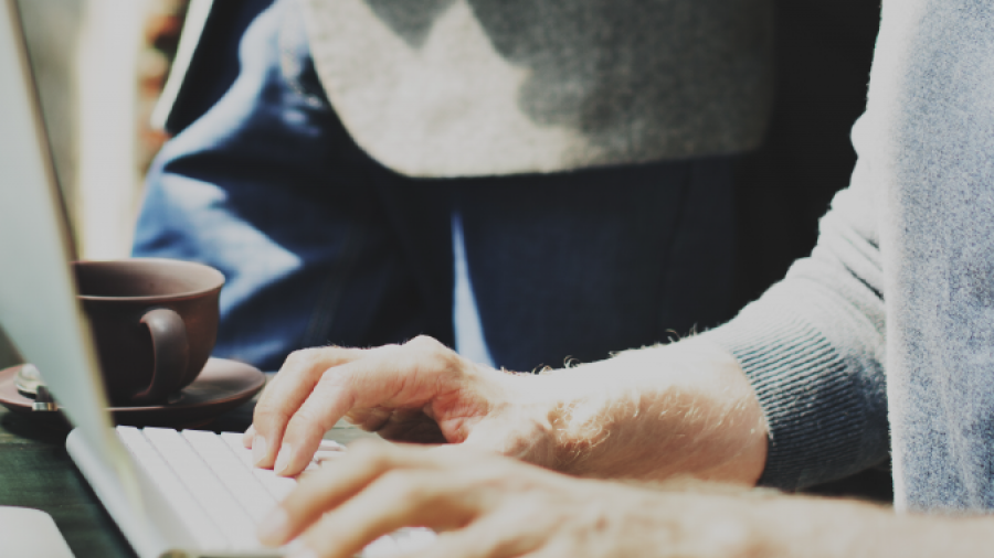 Close-up of a person typing on a keyboard at a desk with a computer monitor, a coffee cup, and another person standing nearby, partially visible in the background.