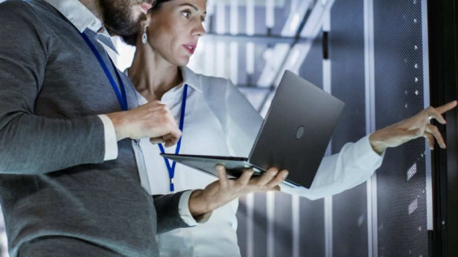 Two people in business attire, wearing lanyards, stand in a server room. One holds a laptop while the other points at a server rack, both focused and engaged in work-related discussion.