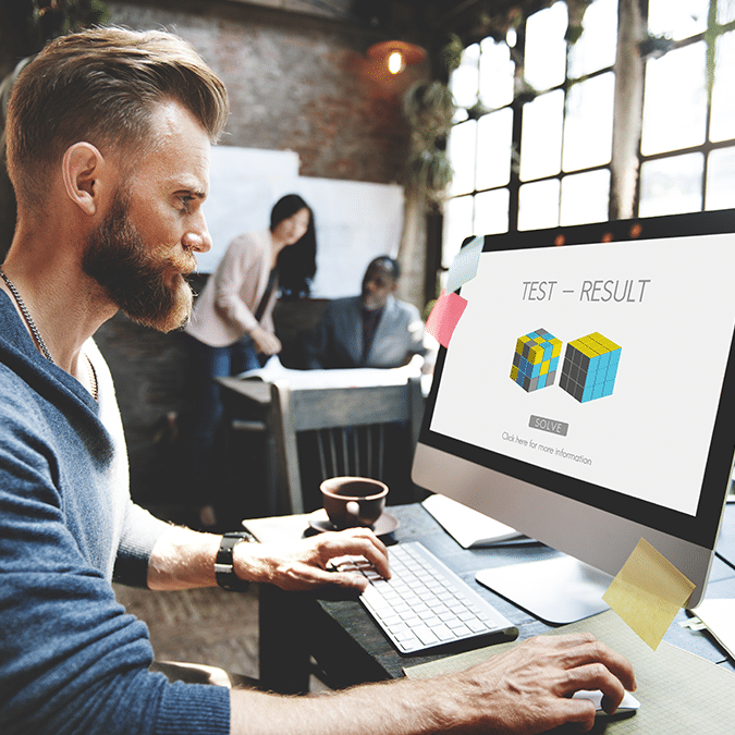 A man with a beard works on a desktop computer in a modern office. His screen displays a test result with images of colored cubes. Two colleagues are blurred in the background at a table.