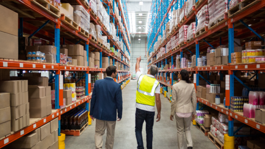 Three people walk down a wide aisle in a large warehouse filled with tall shelves stacked with boxes and goods. One person wears a fluorescent safety vest; the others wear business attire.