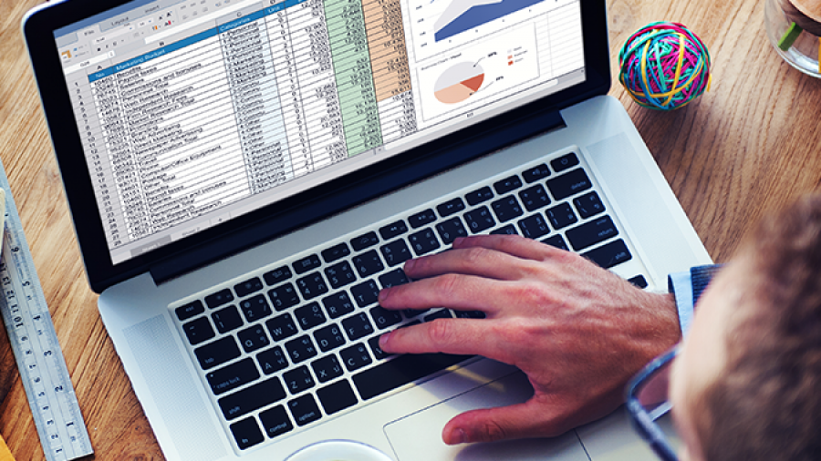 A person holding a cup of coffee works on a laptop displaying a spreadsheet with data tables, charts, and graphs. Office supplies, a notebook, and a colorful rubber band ball are on the wooden desk.