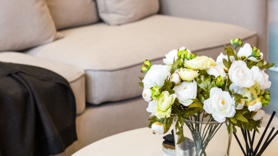 A modern living room scene with a beige sofa, a dark throw blanket, and a round white coffee table. On the table is a glass vase filled with white and yellow flowers, and a silver tray holding decor and reed diffusers.
