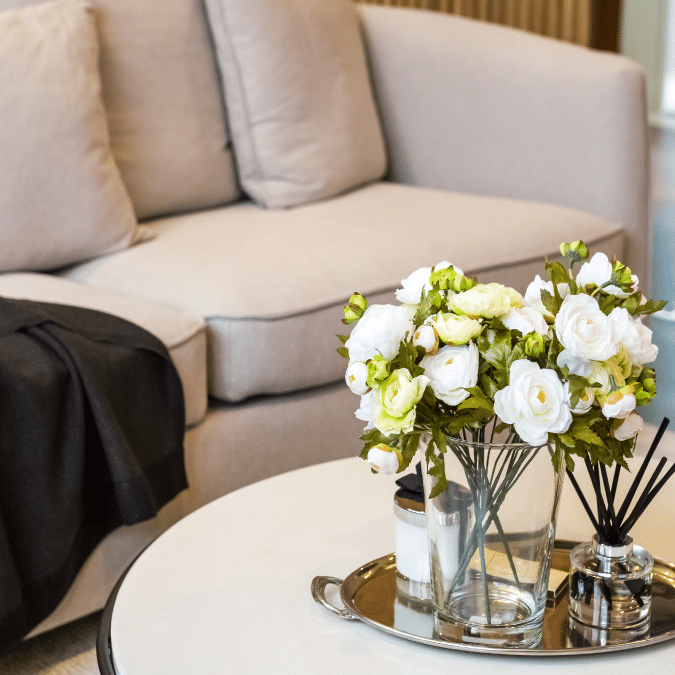 A modern living room scene with a beige sofa, a dark throw blanket, and a round white coffee table. On the table is a glass vase filled with white and yellow flowers, and a silver tray holding decor and reed diffusers.