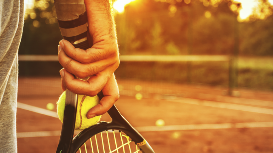 A person stands on a clay tennis court at sunset, holding a tennis racket and a yellow ball. Only their hand, racket, and part of their shorts are visible; tennis balls are scattered on the court.