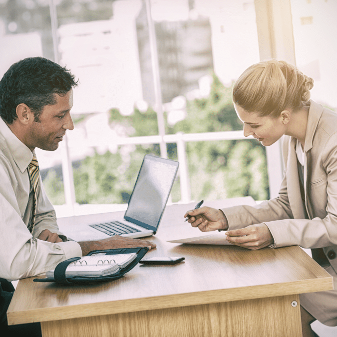 Two business professionals sit across a desk in an office, discussing documents. A laptop, notebook, and smartphone are on the desk. Both are focused, and the room is bright with large windows in the background.