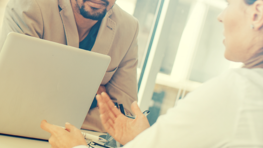 A man in a suit jacket sits at a desk with a laptop, attentively listening to a woman across from him who gestures with her hands as they have a conversation in a bright office setting.