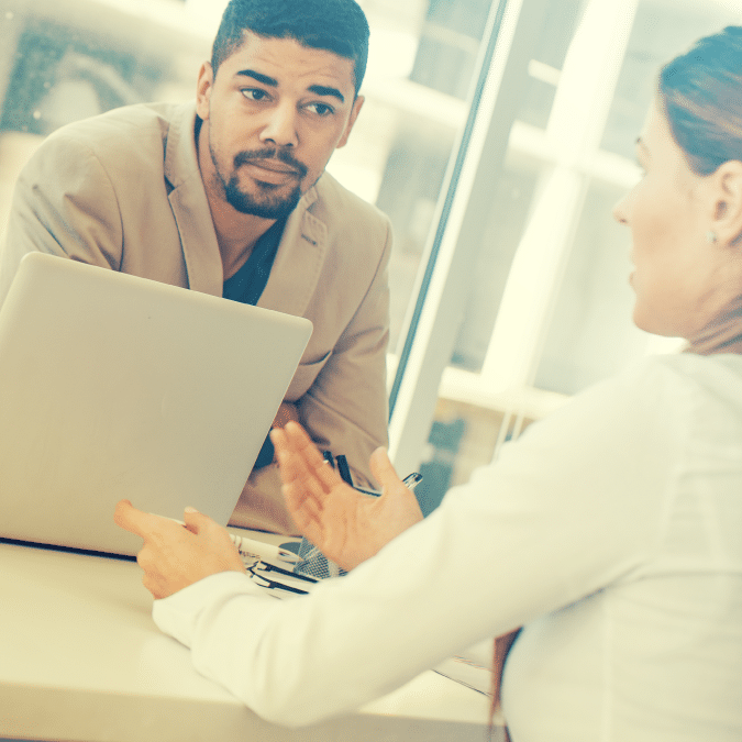 A man in a suit jacket sits at a desk with a laptop, attentively listening to a woman across from him who gestures with her hands as they have a conversation in a bright office setting.
