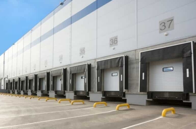 A row of loading docks with closed bay doors at a large industrial warehouse. Each dock is numbered, and protective barriers are placed in front. The building exterior is grey and white under a clear blue sky.