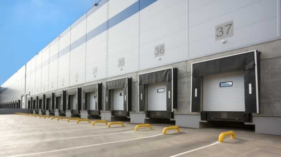 A row of loading docks with closed bay doors at a large industrial warehouse. Each dock is numbered, and protective barriers are placed in front. The building exterior is grey and white under a clear blue sky.
