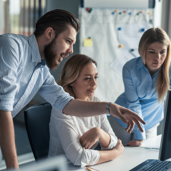 Three colleagues are gathered around a computer in an office, with one man pointing at the monitor and two women watching attentively. A flipchart with papers and notes is visible in the background.