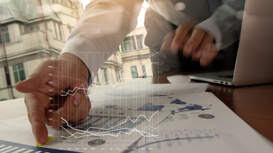 A person points at printed charts and graphs on a desk beside a laptop, with a transparent digital graph overlay, in a well-lit office with large windows.