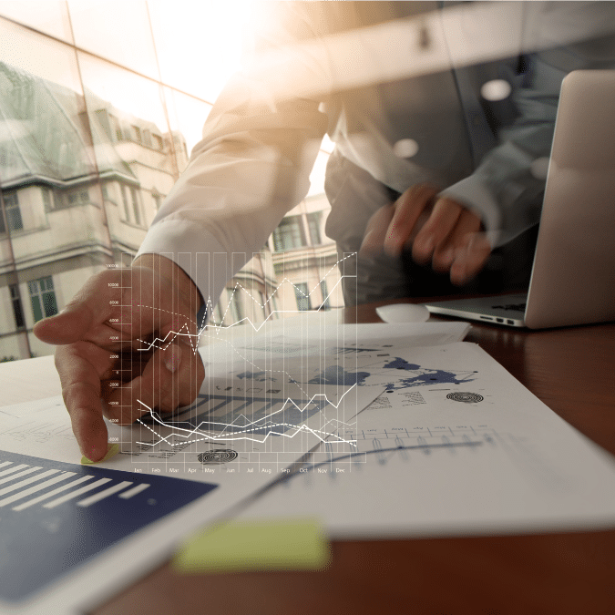 A person points at printed charts and graphs on a desk beside a laptop, with a transparent digital graph overlay, in a well-lit office with large windows.