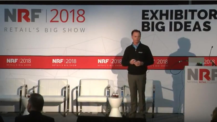 A speaker stands on stage at the NRF 2018 Retails Big Show event, next to three white chairs. The backdrop displays EXHIBITOR BIG IDEAS and NRF 2018.