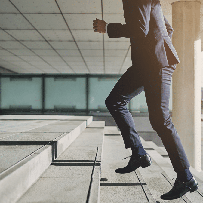 A person in a business suit walks up a set of outdoor concrete stairs, with only their lower body visible. The background features modern architecture and large windows.