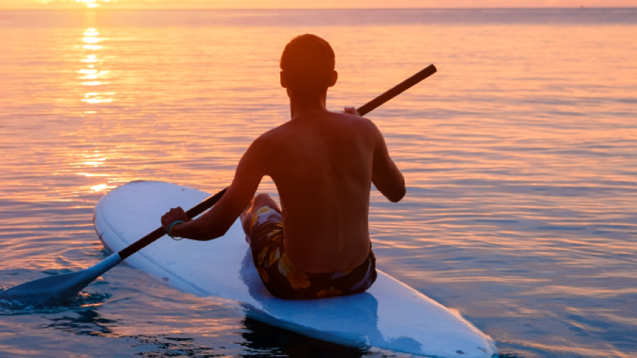 A person paddles on a stand-up paddleboard in calm ocean water at sunset, with vibrant orange and pink hues in the sky and the sun low on the horizon.