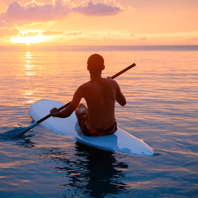 A person paddles on a stand-up paddleboard in calm ocean water at sunset, with vibrant orange and pink hues in the sky and the sun low on the horizon.