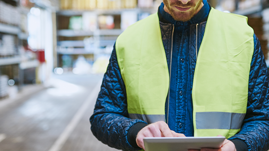 A man wearing a yellow safety vest uses a digital tablet in a warehouse aisle, focusing on the screen with shelves and industrial lighting in the background.