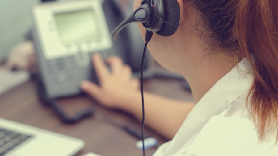 A woman wearing a headset works at a desk, holding a pen and touching a desk phone, with a laptop and notepad in front of her.