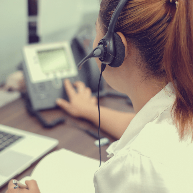 A woman wearing a headset works at a desk, holding a pen and touching a desk phone, with a laptop and notepad in front of her.
