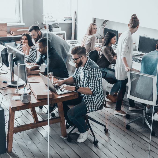 A group of young professionals working together in a modern open-plan office, using computers and collaborating on projects around desks with natural light coming through large windows.