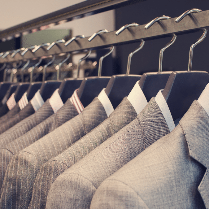 A row of neatly hung mens suit jackets in various shades of grey and patterns on metal hangers, displayed on a rack in a clothing store.