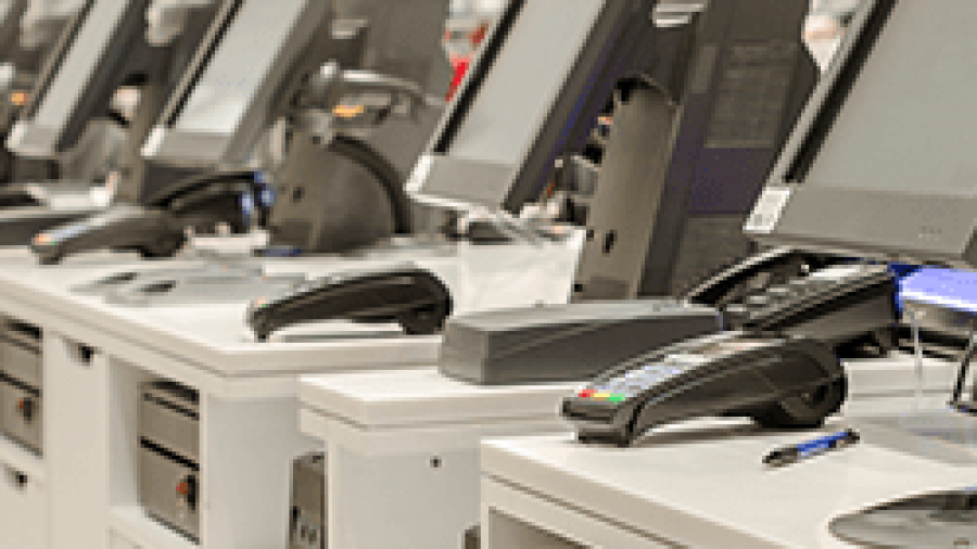 A row of empty retail checkout counters with touchscreen monitors, payment terminals, receipt printers, and pens, in a bright store environment.