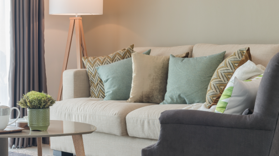 A modern living room with a beige sofa decorated with green and patterned pillows, a gray armchair, a round glass coffee table with books and a plant, and a floor lamp beside closed curtains.