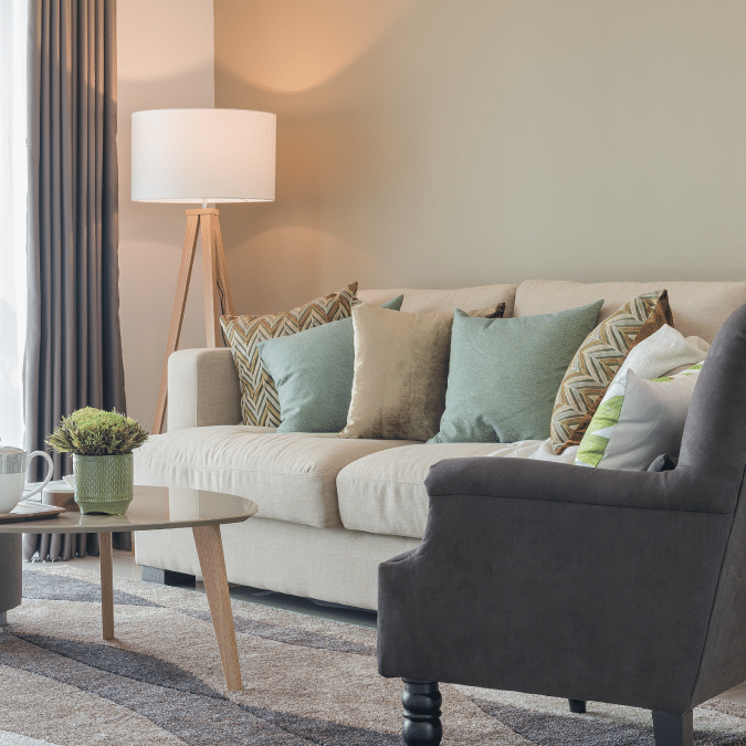 A modern living room with a beige sofa decorated with green and patterned pillows, a gray armchair, a round glass coffee table with books and a plant, and a floor lamp beside closed curtains.