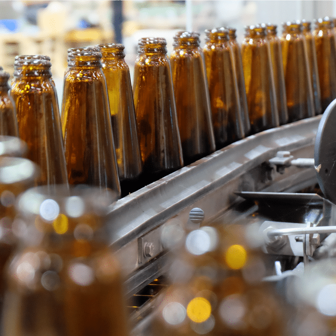 A row of empty brown glass bottles moves along a conveyor belt in a factory setting, ready for filling and packaging.