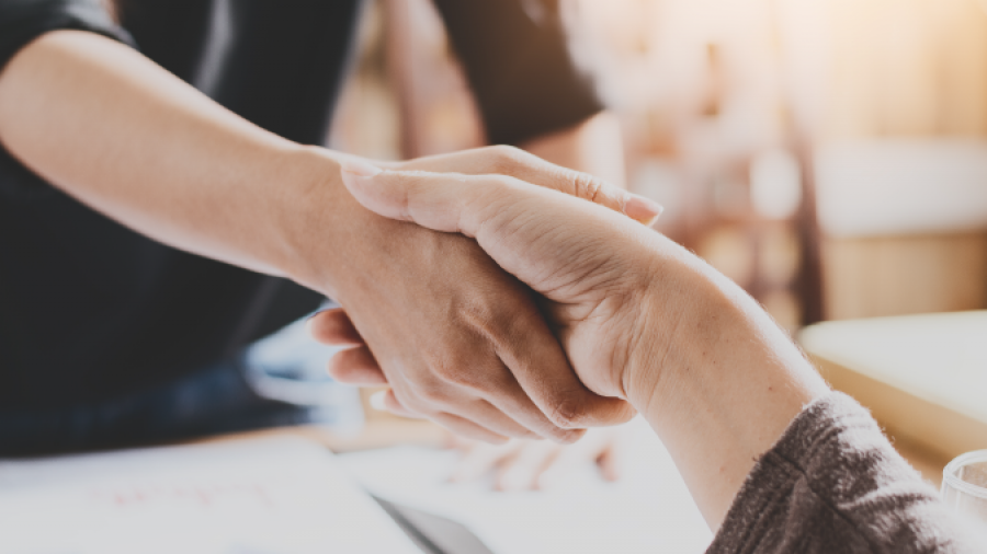 Two people shake hands across a desk with paperwork and a pen, suggesting a successful business agreement or partnership in a professional setting with warm lighting.