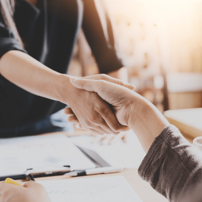 Two people shake hands across a desk with paperwork and a pen, suggesting a successful business agreement or partnership in a professional setting with warm lighting.