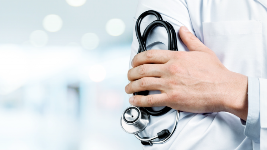 A doctor in a white coat stands with arms crossed, holding a stethoscope, in a bright, blurred medical or hospital setting.
