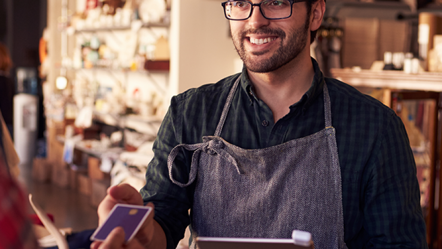 A smiling man wearing glasses and an apron holds a tablet while assisting a customer making a payment with a credit card in a cozy, well-lit shop.