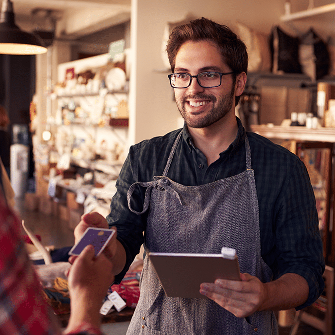 A smiling man wearing glasses and an apron holds a tablet while assisting a customer making a payment with a credit card in a cozy, well-lit shop.