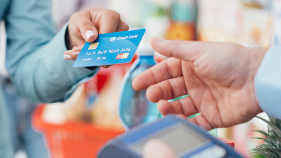 A person holds out a blue credit card to make a payment using a card reader held by another person in a grocery store setting, with a shopping cart visible in the background.
