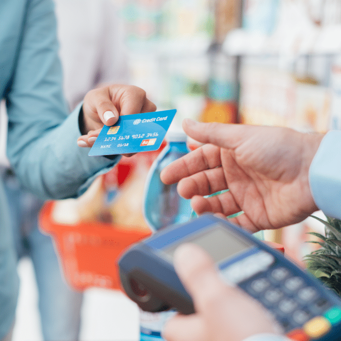 A person holds out a blue credit card to make a payment using a card reader held by another person in a grocery store setting, with a shopping cart visible in the background.