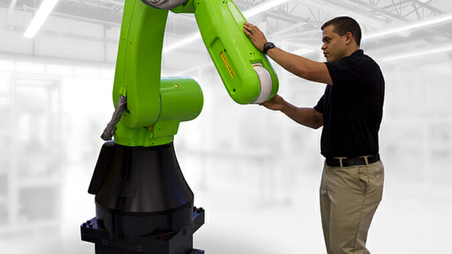 A man in a black shirt and khaki pants interacts with a large green industrial robotic arm inside a bright, modern factory, showcasing innovative design build services in action.