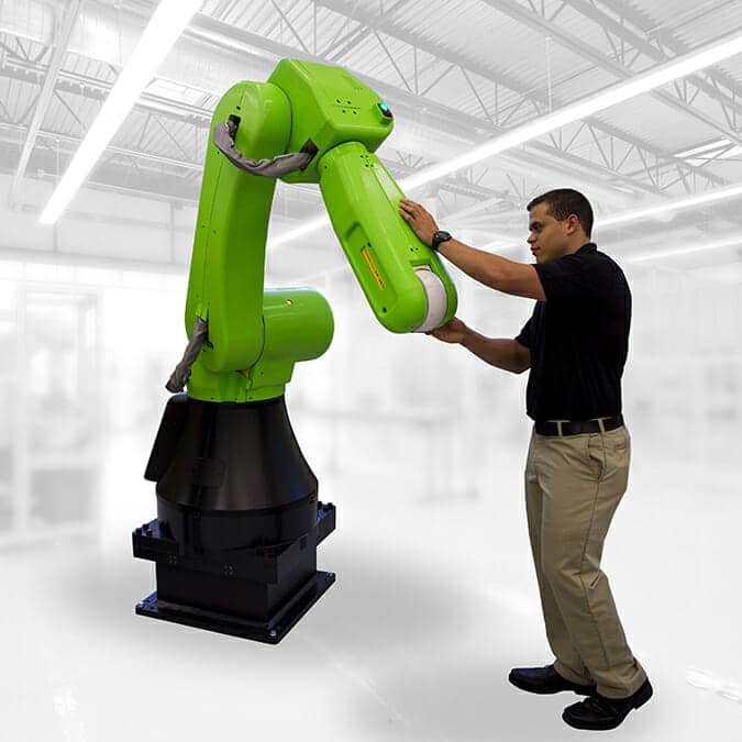 A man in a black shirt and khaki pants interacts with a large green industrial robotic arm inside a bright, modern factory, showcasing innovative design build services in action.
