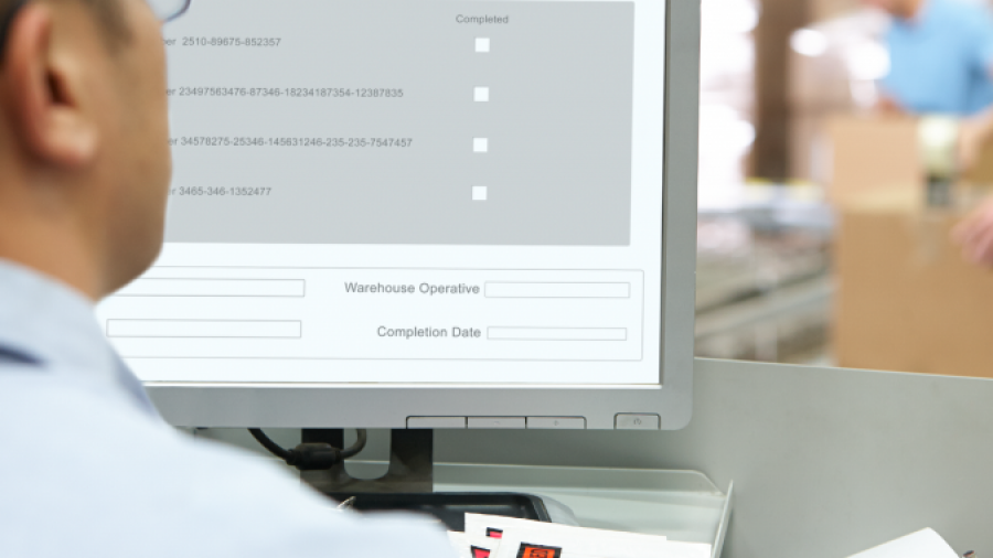A person wearing glasses sits at a desk using a computer with a warehouse management interface on the screen; shelves and another worker are seen blurred in the background.